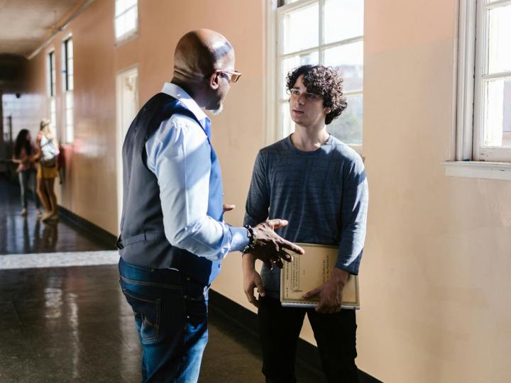 a young man talking to his professor while standing on a hallway